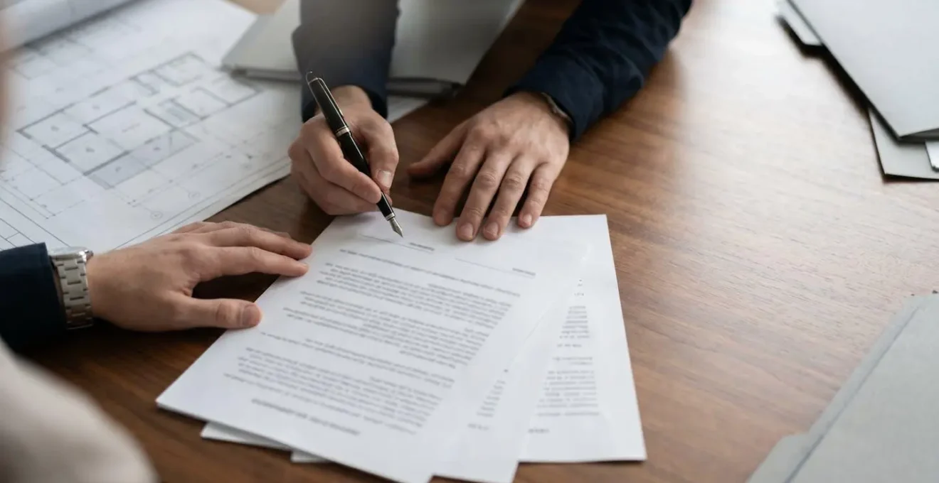 Scène professionnelle de signature de documents immobiliers sur un bureau en bois avec stylo et dossiers